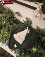 Load image into Gallery viewer, A green bandana with a gold embroidered tree and a pink bow on top, displayed on a white surface surrounded by greenery.